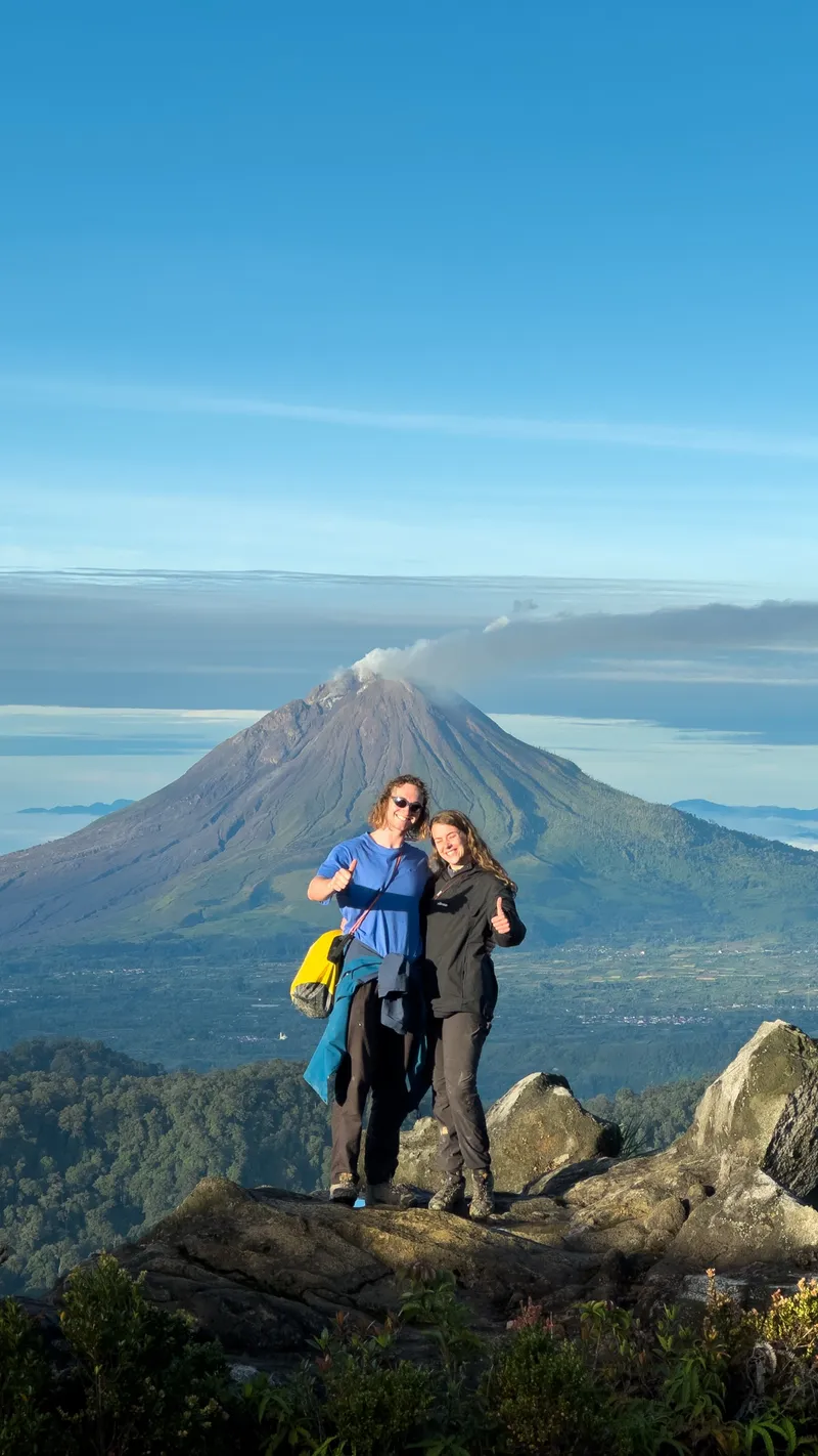 Two guests giving a thumbs-up on the summit rocks with Mount Sinabung behind