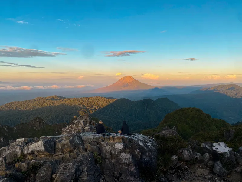 Two hikers sitting on the summit cliff as first light hits Mount Sinabung
