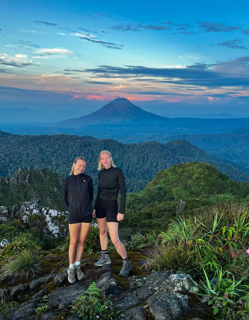 Two guests smiling at dawn on the Mount Sibayak summit with Mount Sinabung behind